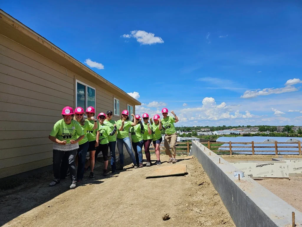 A group of enthusiastic volunteers wearing green “Women Build” t-shirts and pink hard hats pose and smile in front of a Habitat for Humanity home under construction.