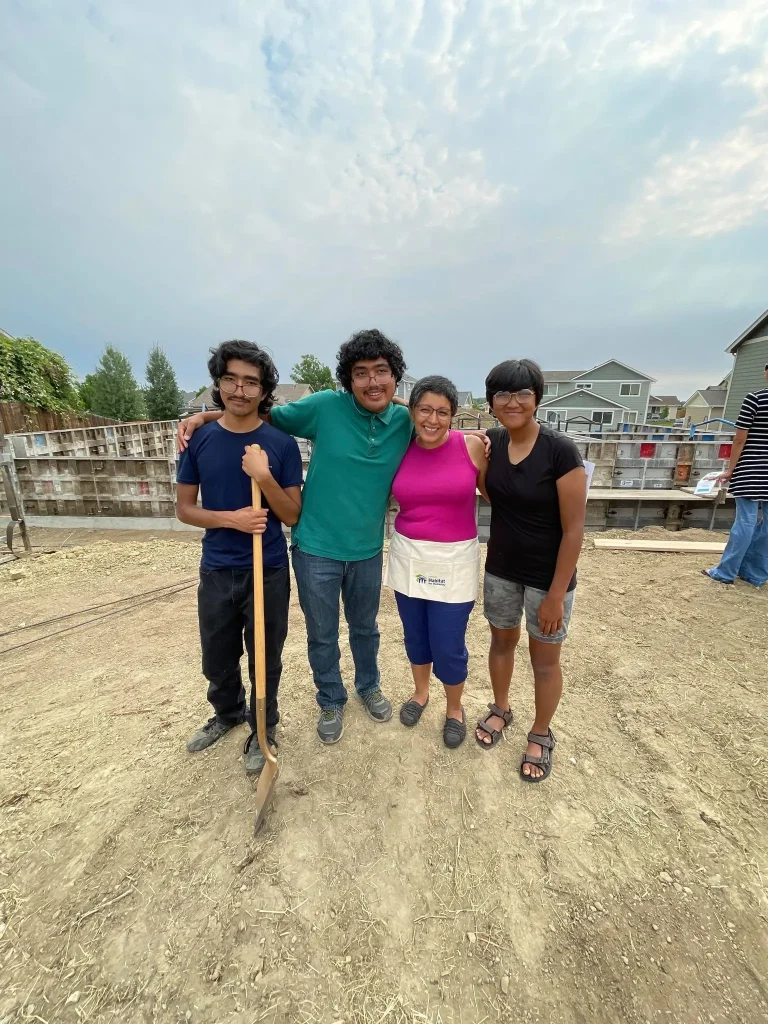 A smiling family of four stands arm-in-arm at a Habitat for Humanity build site, with partially constructed foundations and homes in the background.