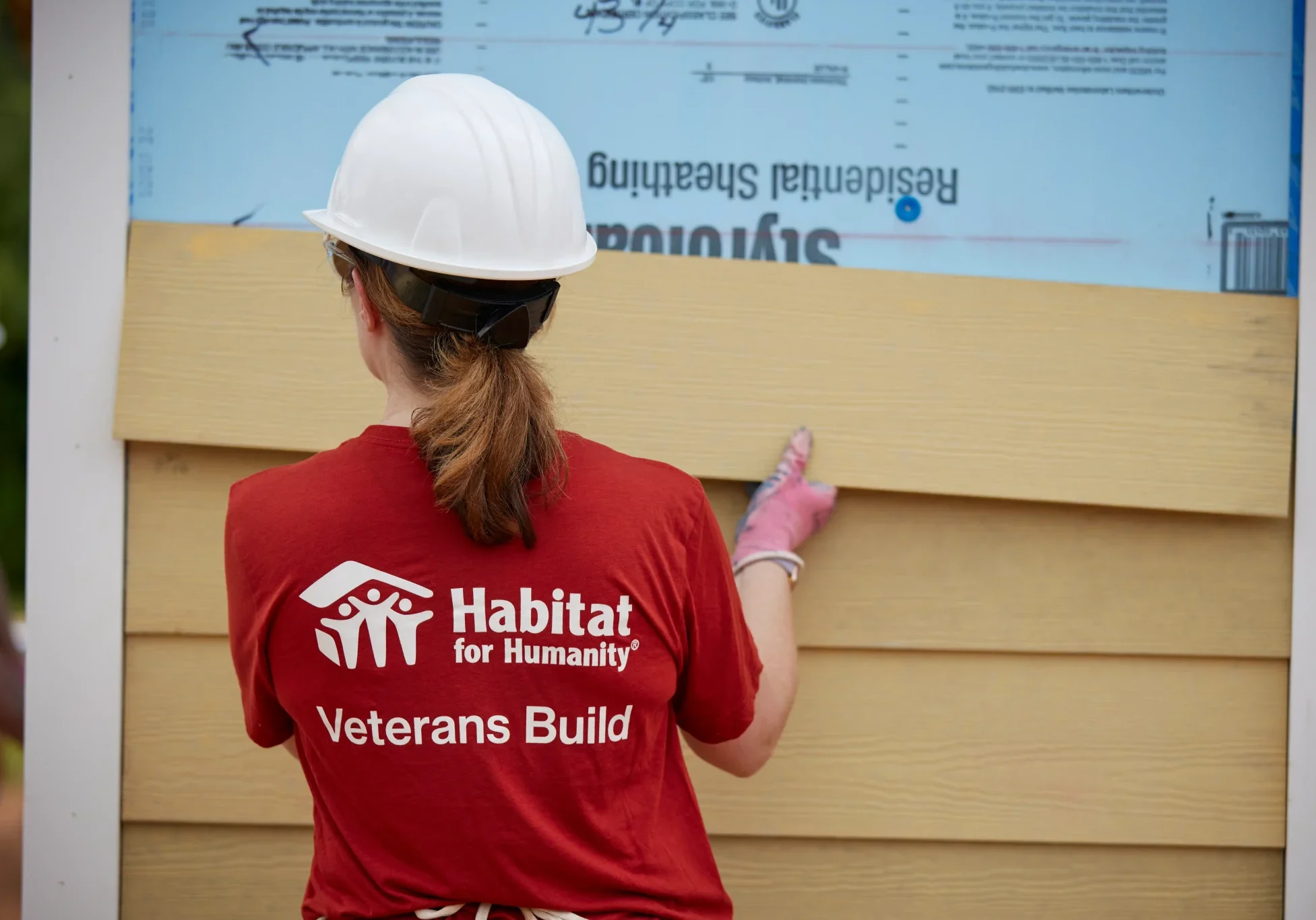 A volunteer in a Veterans Build shirt putting up siding on a house.